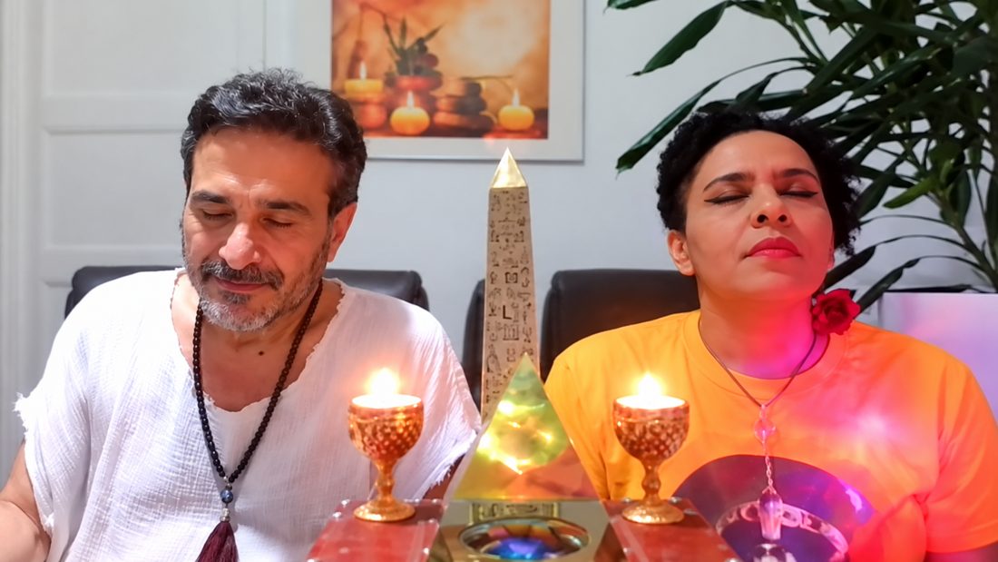 Ayam e Ayala de olhos fechados, em meditação, diante de duas velas acesas e de um obelisco, em um encontro online