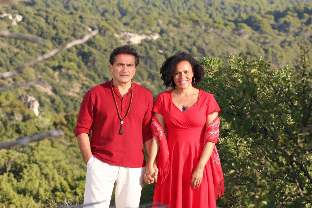 Ayam e Ayala de mãos dadas, vestidos de vermelho, diante de uma encosta de mata